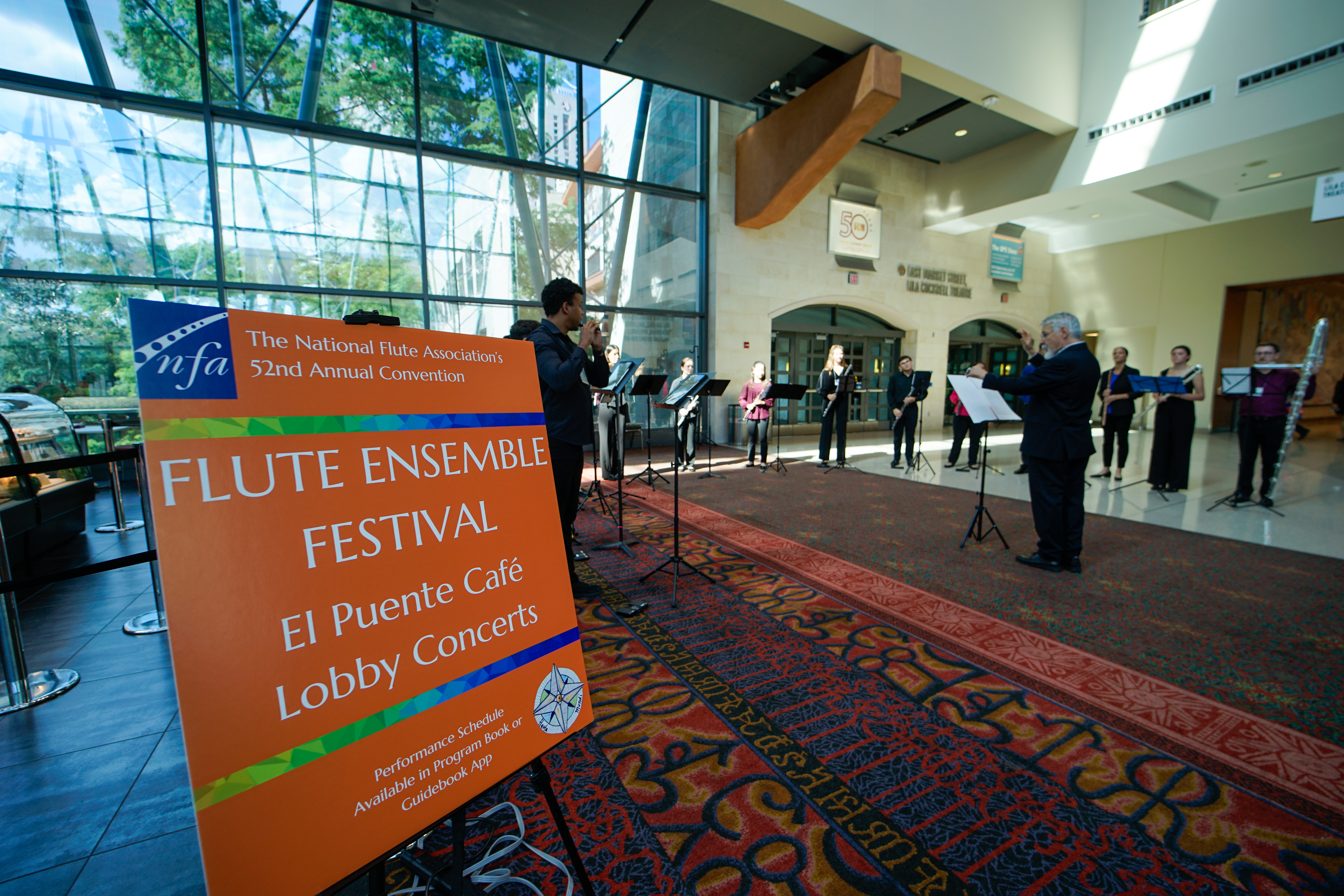 A flute choir performs in a convention center lobby. a sign reads "flute ensemble festival"